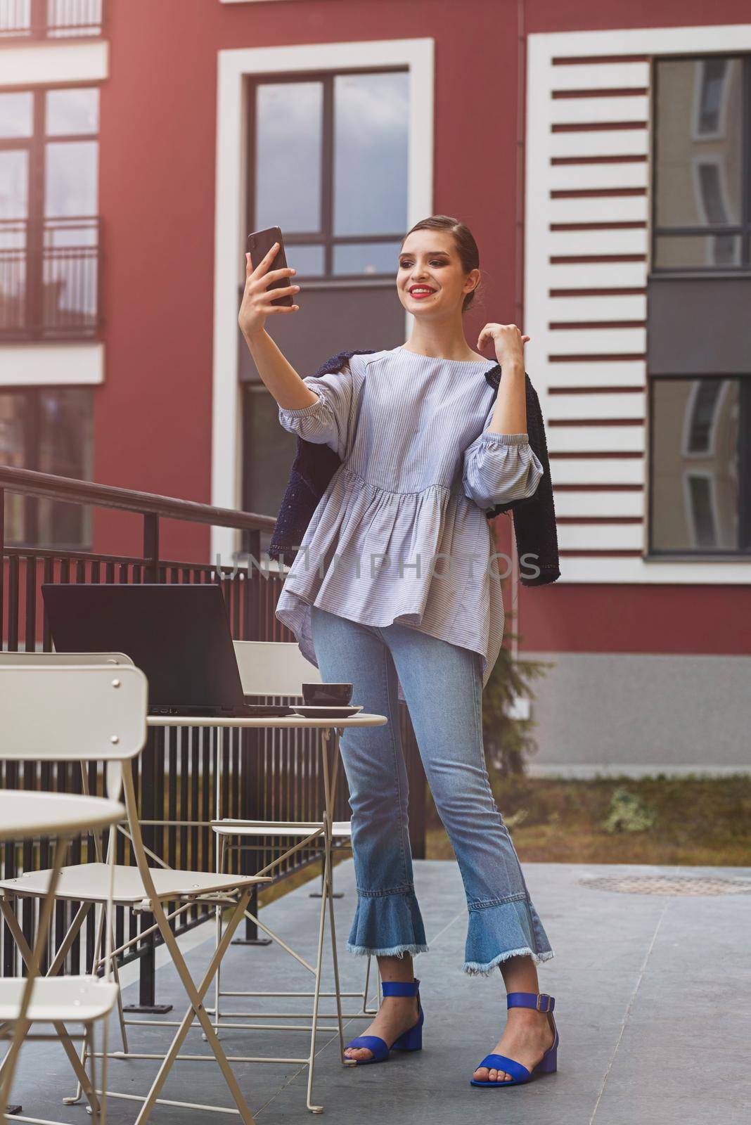 Charming happy woman student communicates by phone and use laptop computer to prepare for the course work. Concept of working outdoors