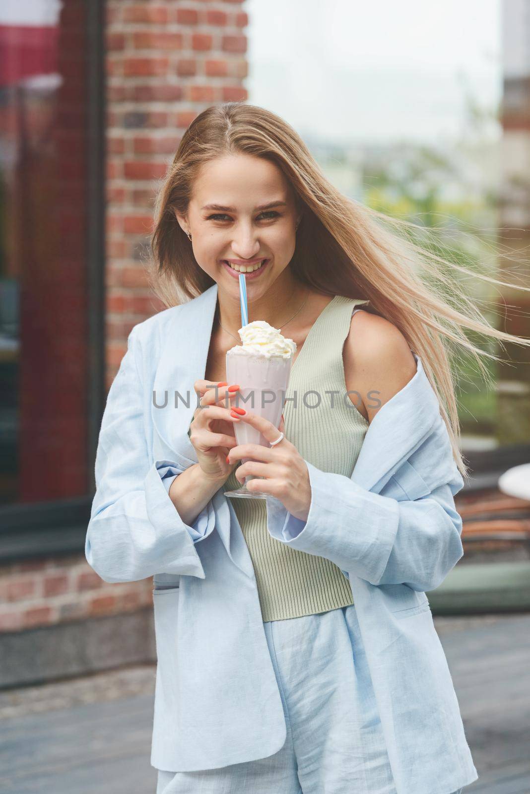 A woman's hand holds a refreshing cocktail in a glass with a tube. tanned woman wearing a beige T-shirt and a blue cotton jacket. Orange manicure