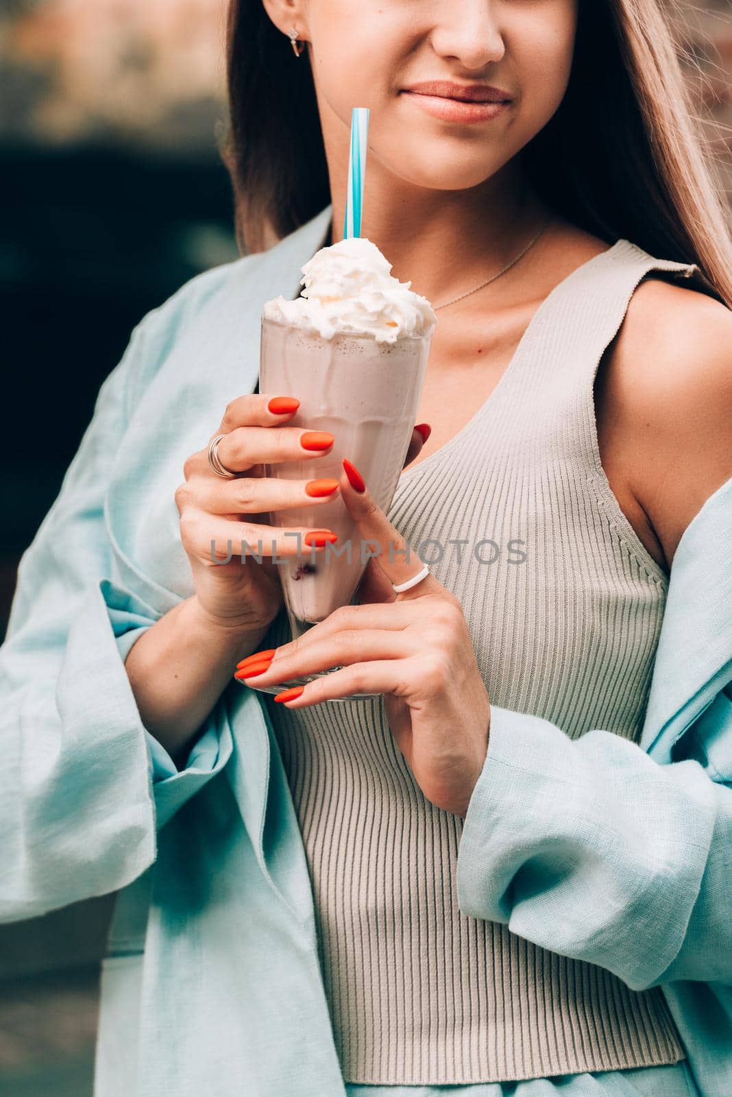 A woman's hand holds a refreshing cocktail in a glass with a tube. tanned woman wearing a beige T-shirt and a blue cotton jacket. Orange manicure