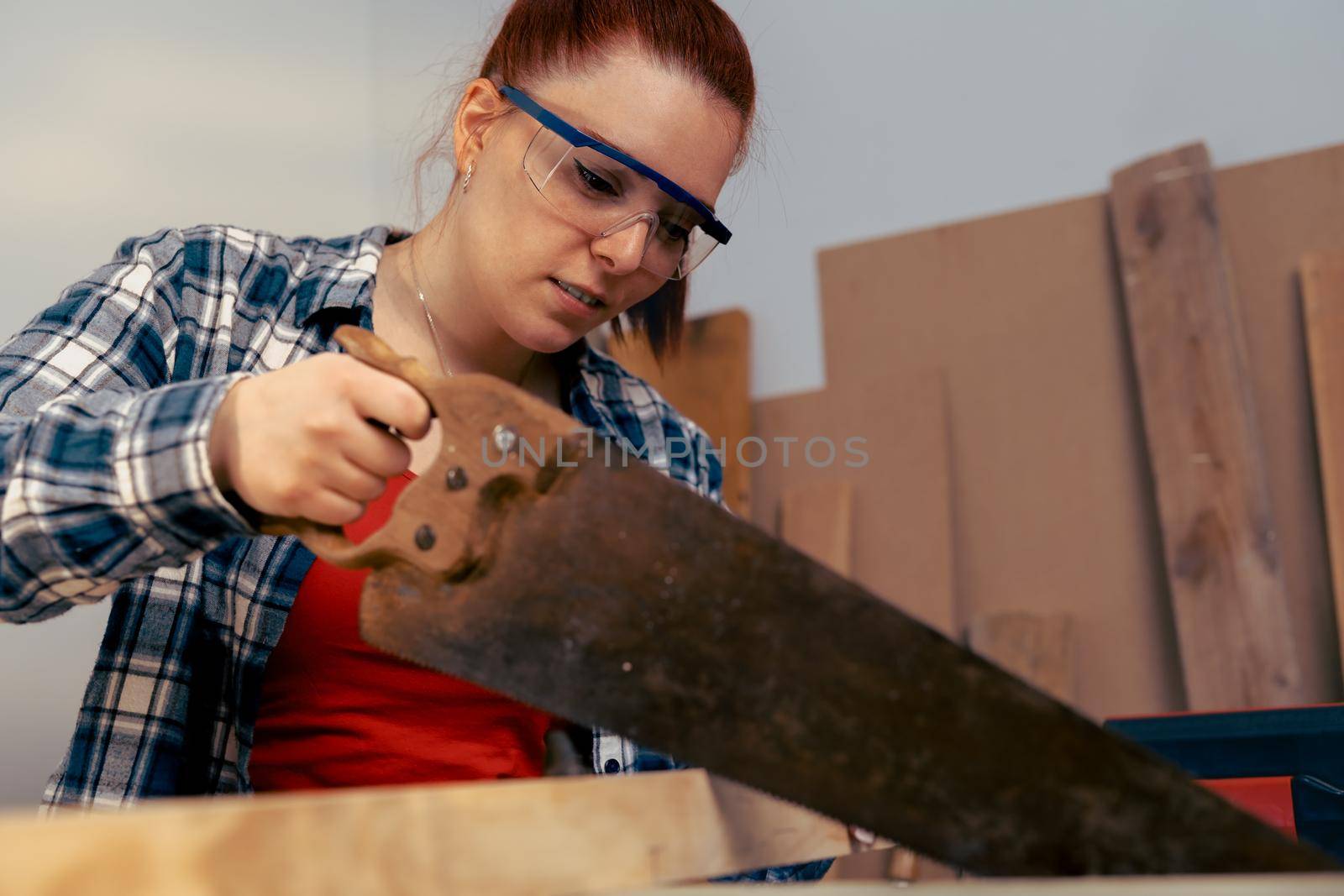 close-up of a young red-haired woman carpenter sawing a wooden board with a saw, in her small carpentry shop. by CatPhotography