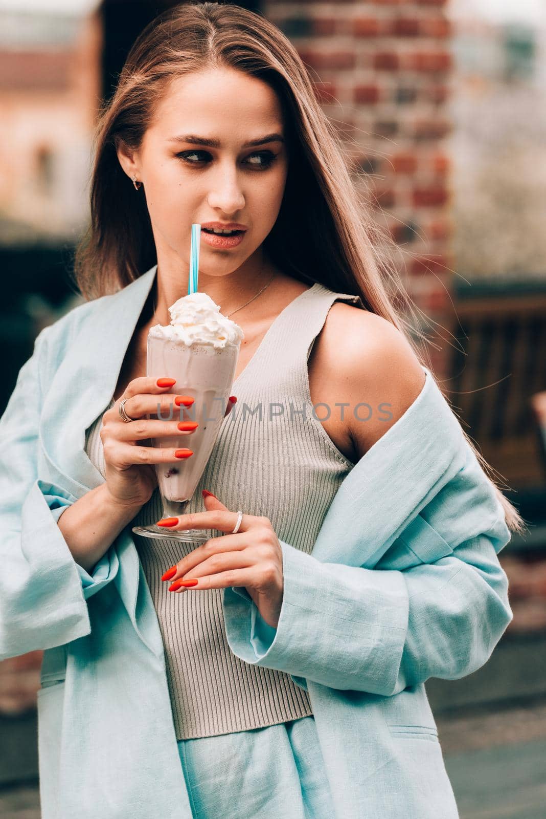 A woman's hand holds a refreshing cocktail in a glass with a tube. tanned woman wearing a beige T-shirt and a blue cotton jacket. Orange manicure