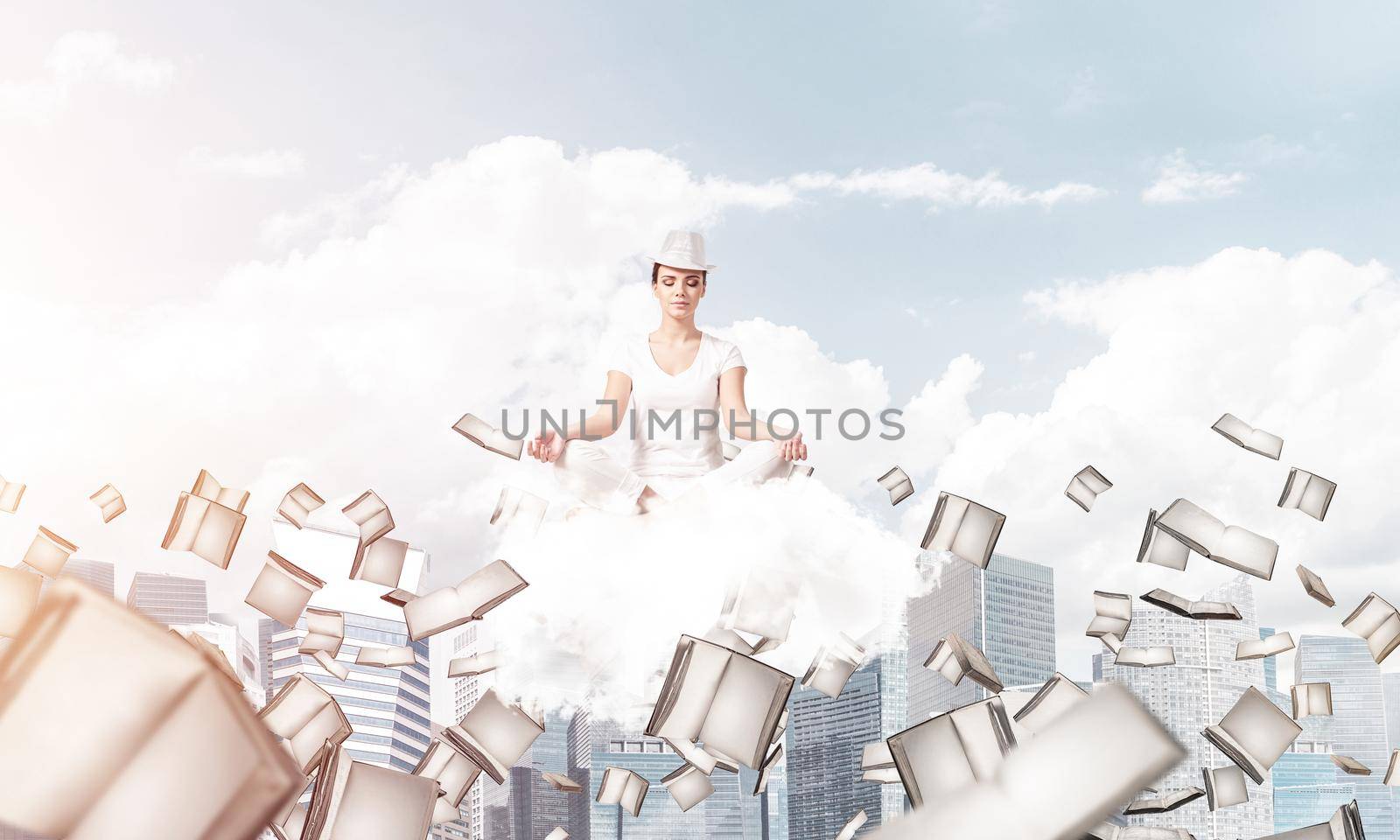 Woman in white clothing keeping eyes closed and looking concentrated while meditating on cloud among flying books with cityscape view on background.