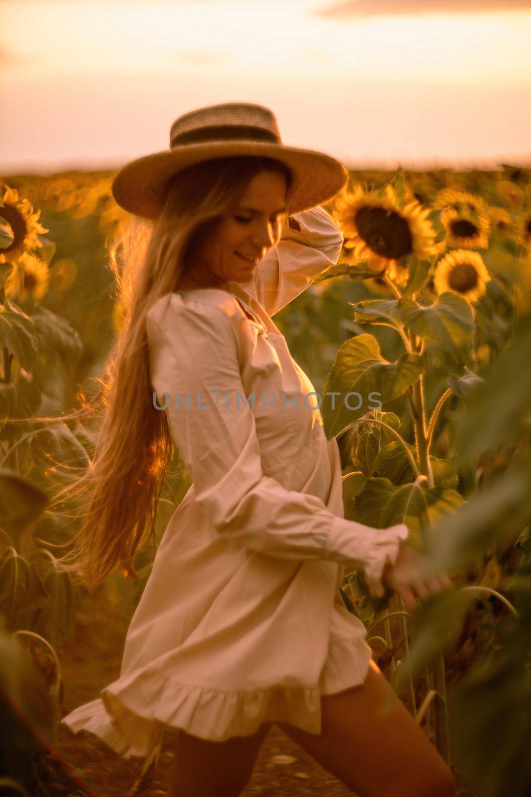 Beautiful middle aged woman looks good in a hat enjoying nature in a field of sunflowers at sunset. Summer. Attractive brunette with long healthy hair