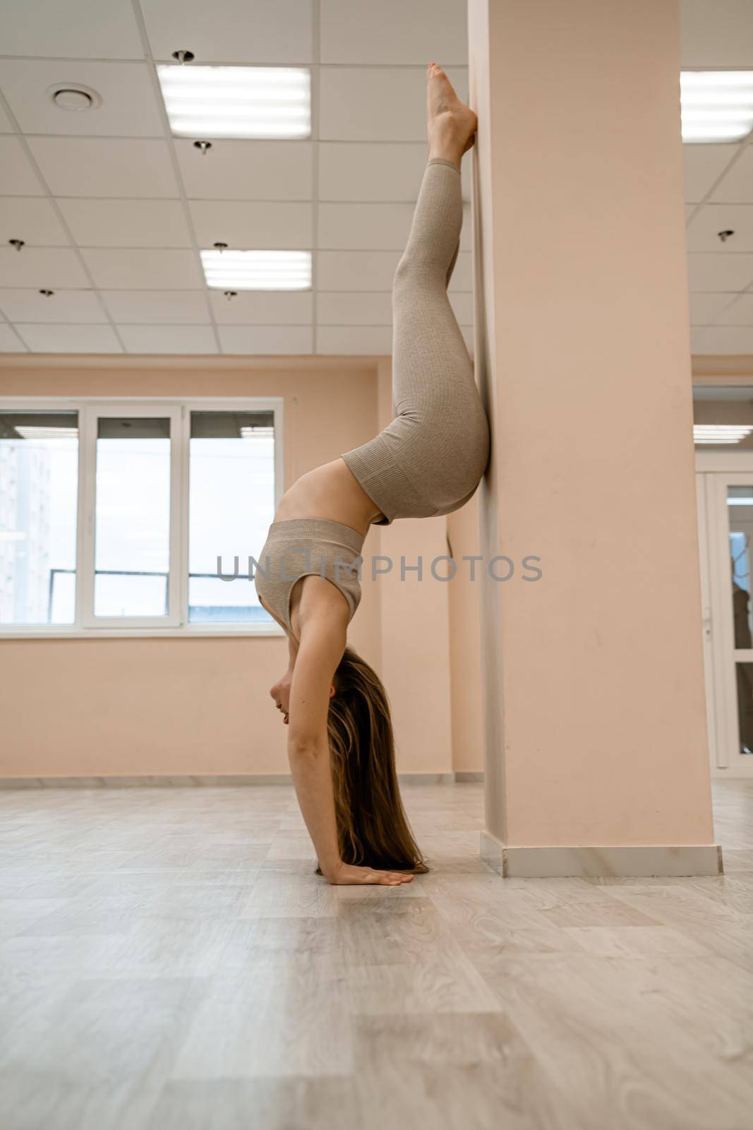 The girl is training in the yoga studio. Dressed in a beige tracksuit doing a handstand against the wall. Healthy lifestyle and yoga concept