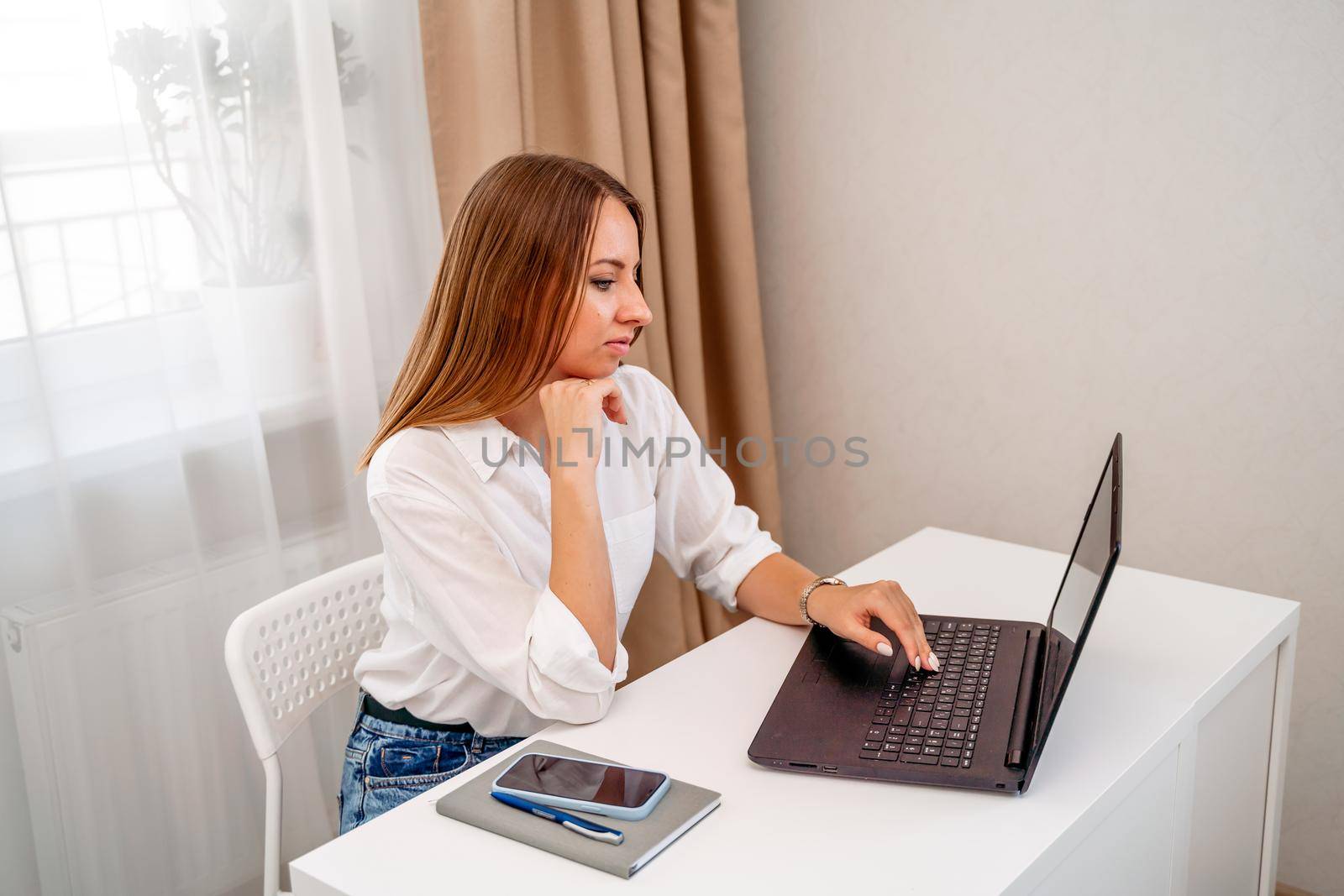 European professional woman is sitting with a laptop at a table in a home office, a positive woman is studying while working on a PC. She is wearing a beige jacket and jeans and is on the phone. by Matiunina