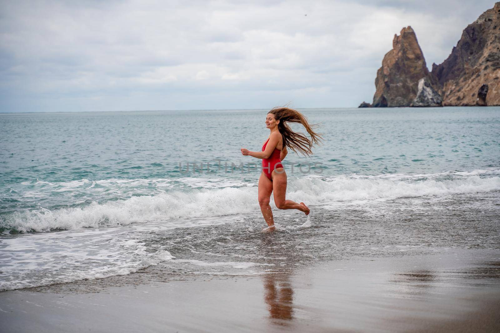 A beautiful and sexy brunette in a red swimsuit on a pebble beach, Running along the shore in the foam of the waves by Matiunina