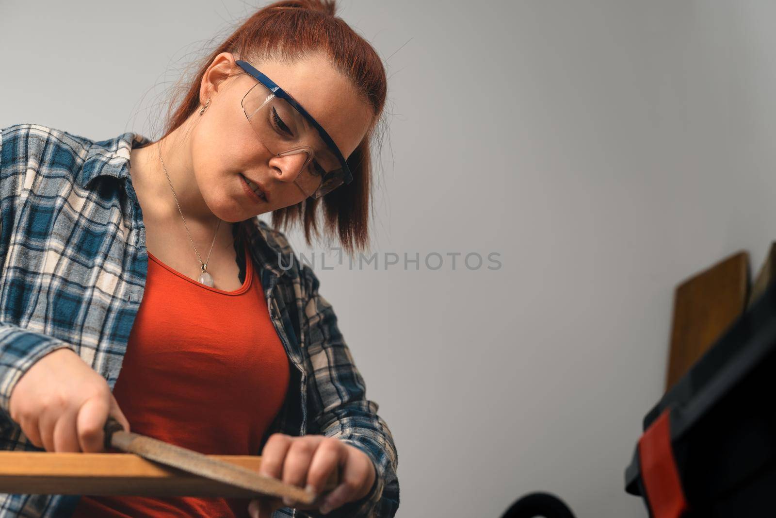 close-up of a young red-haired woman, carpenter filing a wooden board with a file, in her small carpentry shop. by CatPhotography