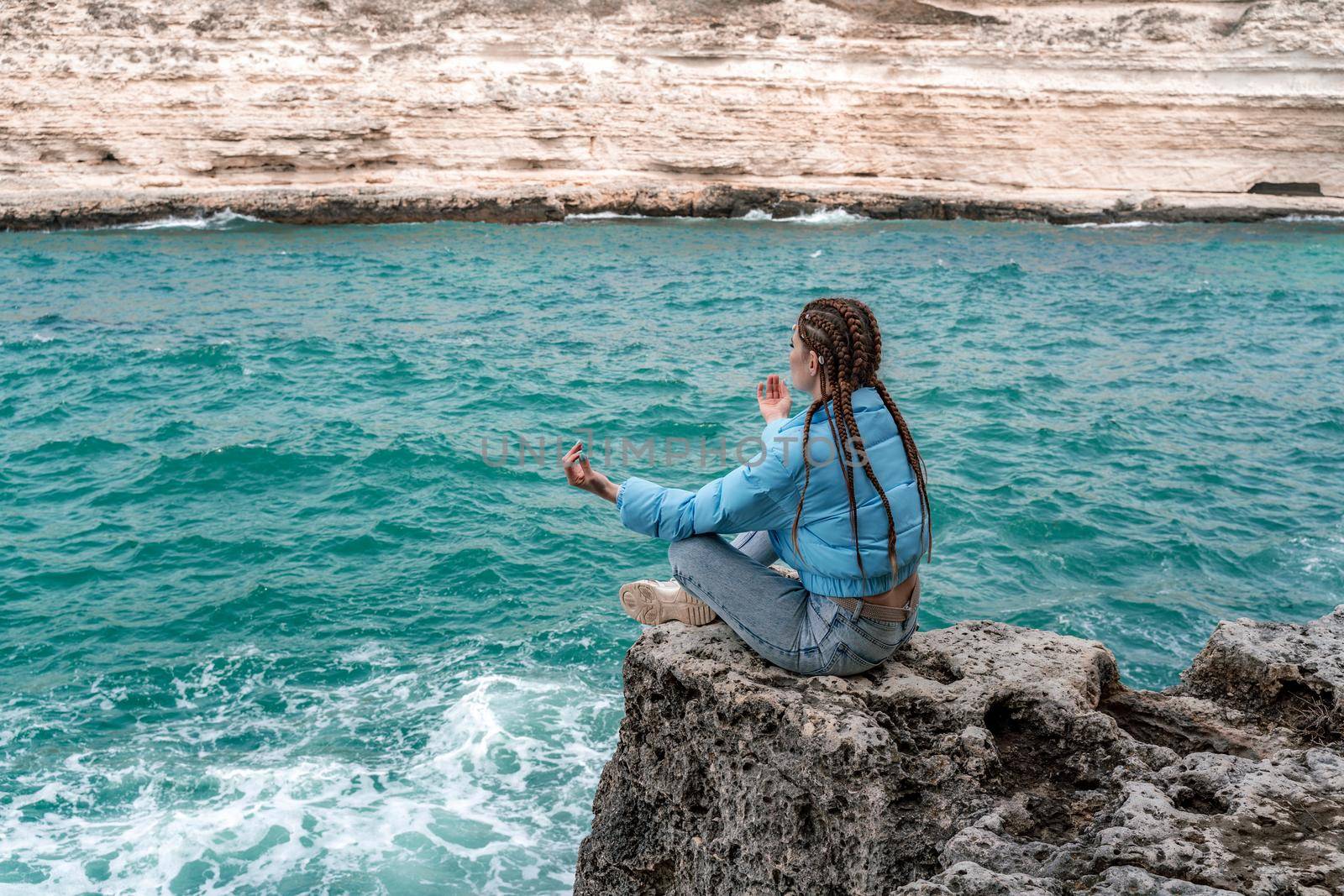 A woman in a blue jacket sits on a rock above a cliff above the sea, looking at the stormy ocean. Girl traveler rests, thinks, dreams, enjoys nature. Peace and calm landscape, windy weather. by Matiunina