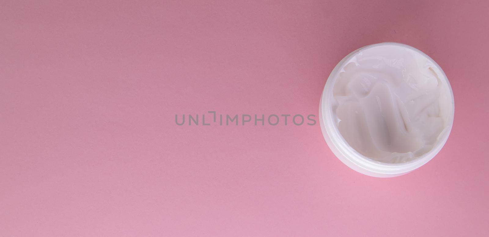 White cosmetic face cream in a round open jar on a pink background, top view, copy of the space on the left.