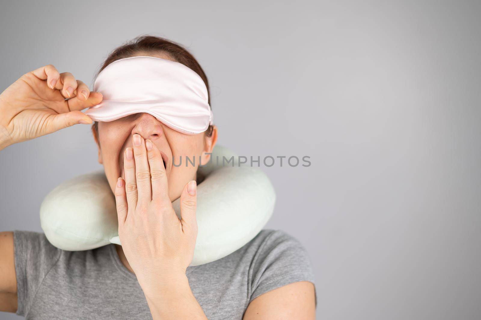 Caucasian woman yawns with travel pillow and sleep mask on white background