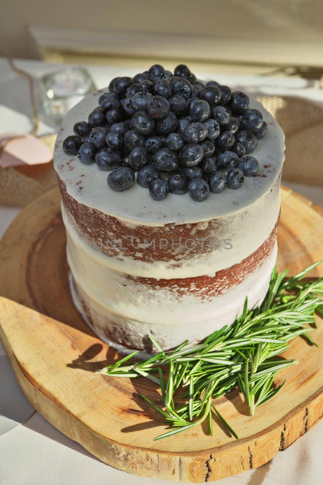 High Angle View of Rustic Homemade Naked Layered Wedding Cake with Vanilla Icing, Fresh Blueberries, and Rosemary, on Wood Slice