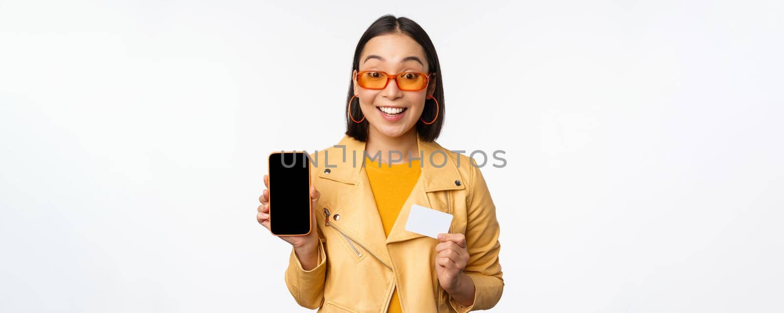 Online shopping and people concept. Stylish asian woman showing mobile phone screen and credit card, smartphone application, standing over white background. Copy space