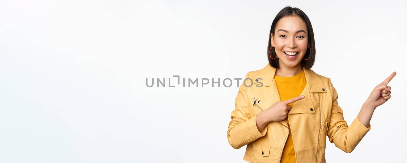 Enthusiastic asian girl pointing fingers right, showing advertisement and smiling, inviting to the store, standing over white background.