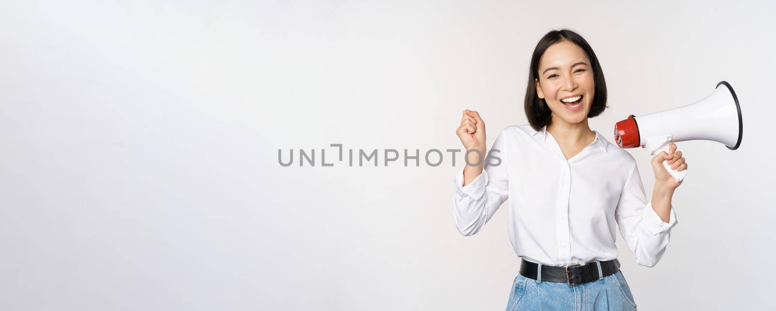 Smiling young asian woman posing with megaphone, concept of news, announcement and information, standing over white background.