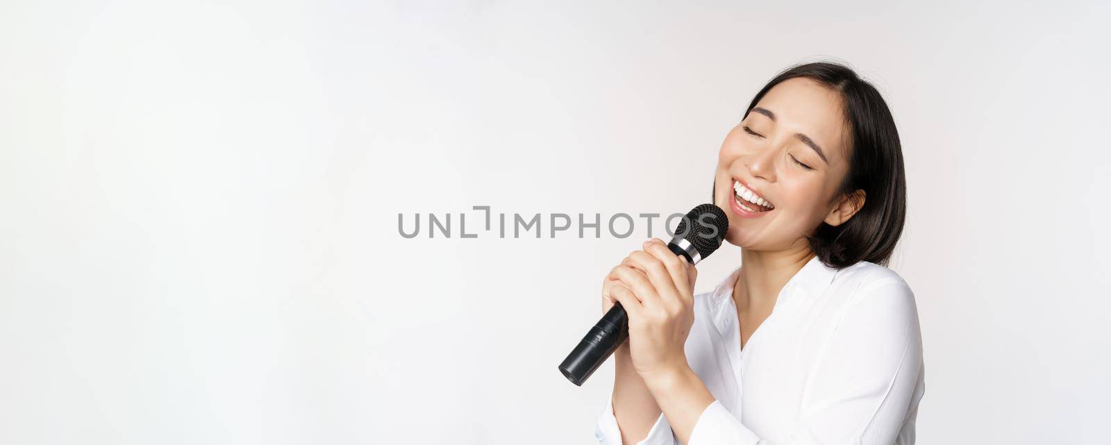 Close up portrait of asian woman singing in microphone at karaoke, standing over white background.