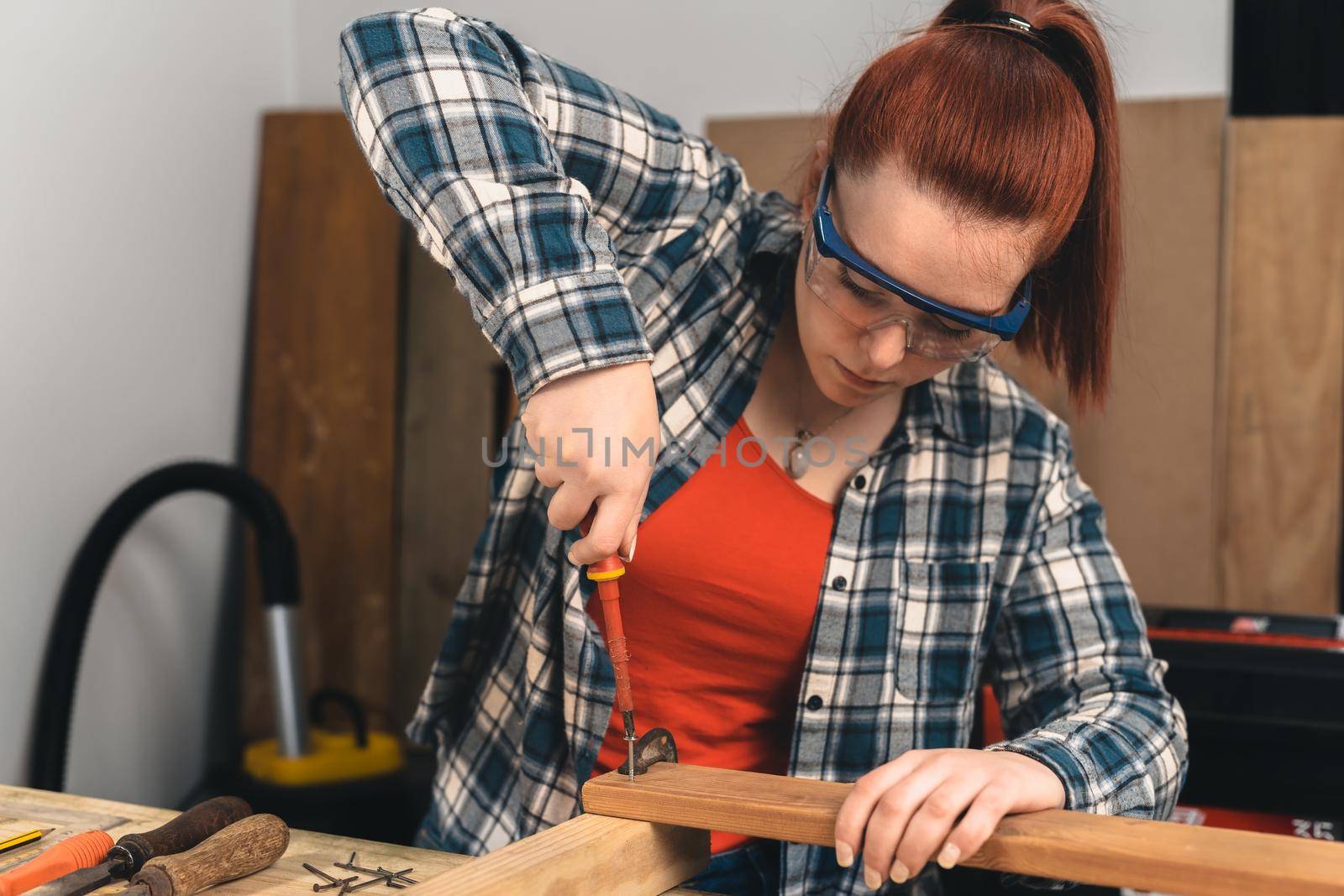 Detail of the hands of a young red-haired carpenter woman, concentrated and precise, working on the design of the wood in a small carpentry workshop, dressed in a blue checked shirt and a red t-shirt. woman carpenter screwing a screw into a wooden plank. Warm light indoors, background with wooden slats. Horizontal.
