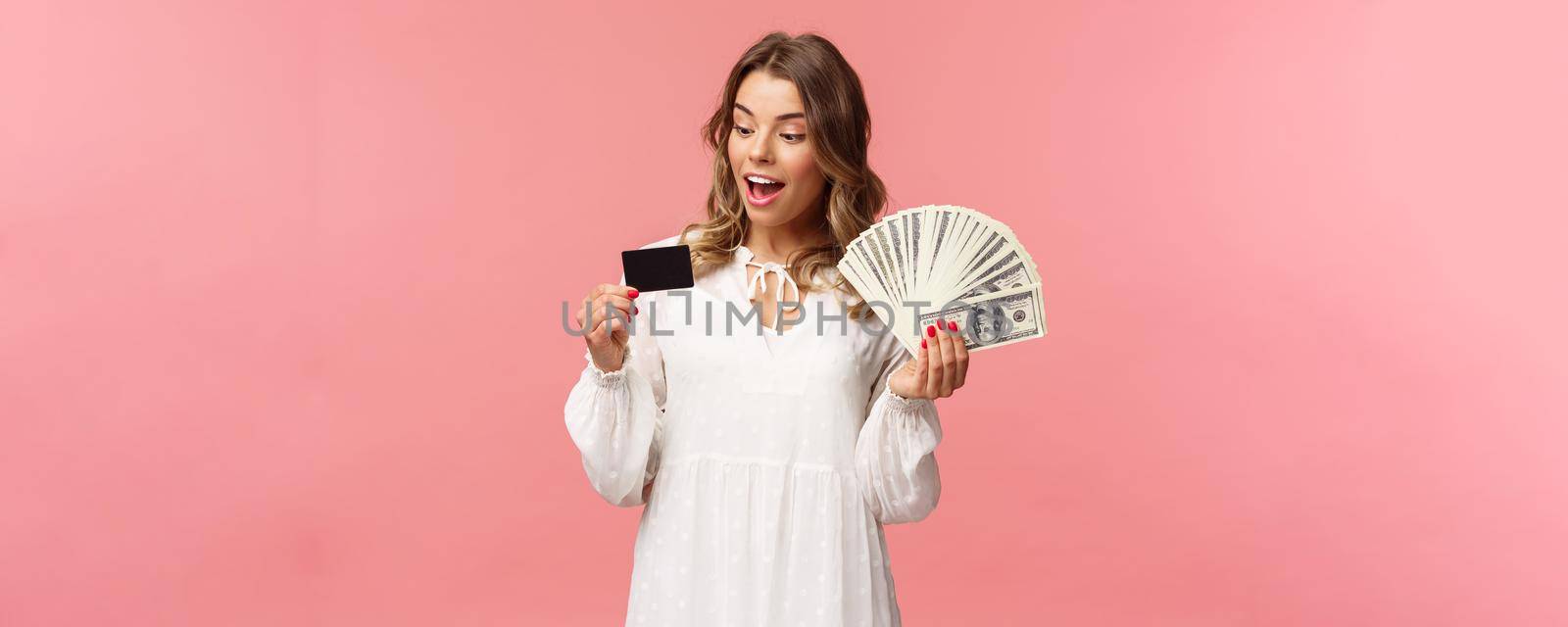 Portrait of rich good-looking blond girl in white dress, holding dollars money and credit card, winning lottery, think how invest, standing happy pink background. Copy space