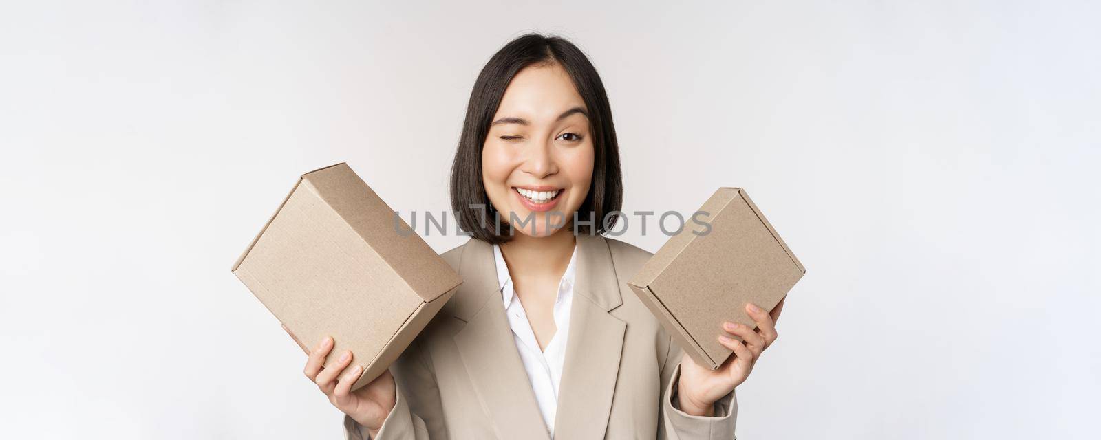 Image of saleswoman, asian businesswoman holding boxes with company brand product, smiling at camera, standing against white background.