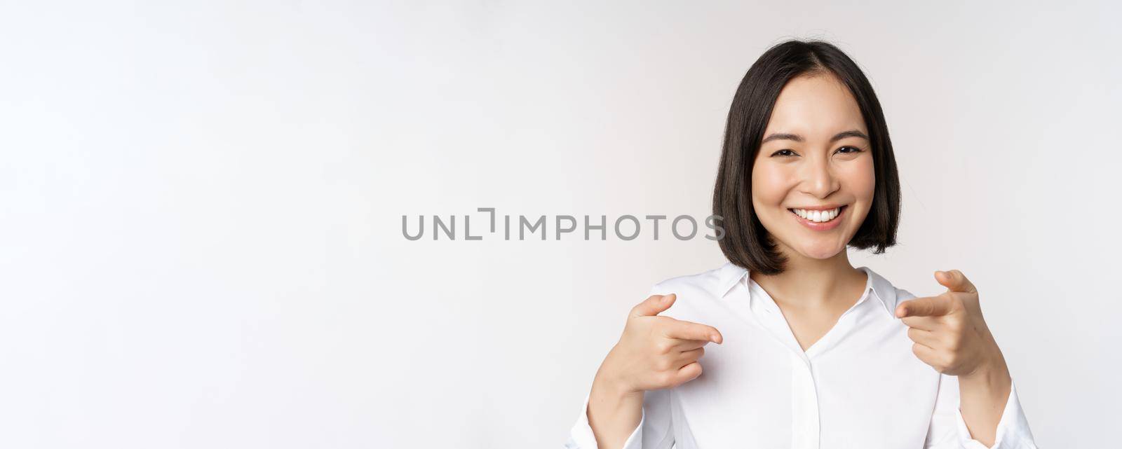 Close up of enthusiastic young woman smiling, pointing fingers at camera, choosing you, inviting and recruiting people, standing over white background.
