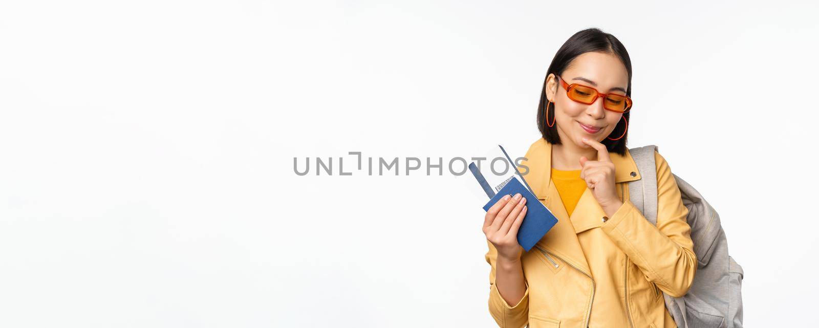 Asian girl tourist with boarding tickets and passport going abroad, holding backpack, thinking of travelling, standing over white background.