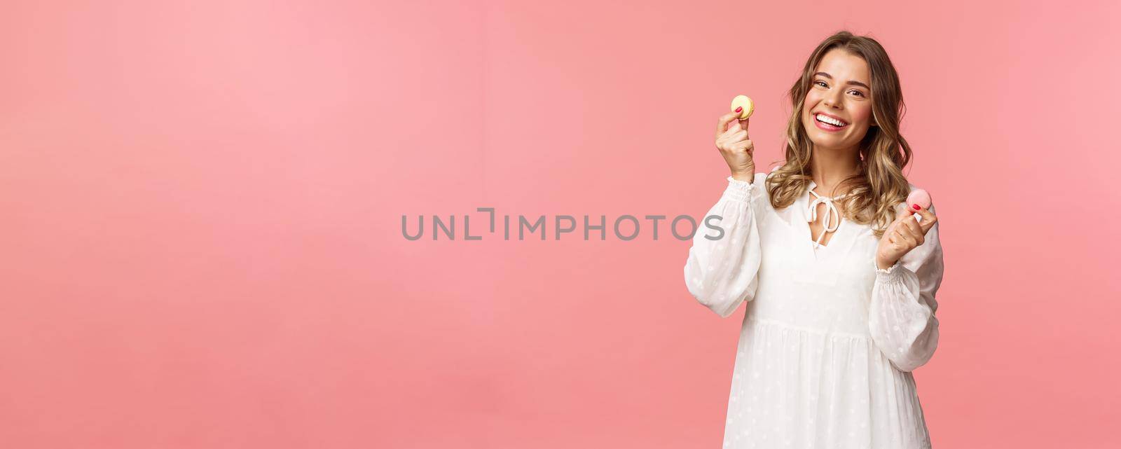 Holidays, spring and party concept. Portrait of cute romantic blond girl in white dress, licking lips as tempting to eat delicious dessert, holding two macarons and look pleased, pink background.