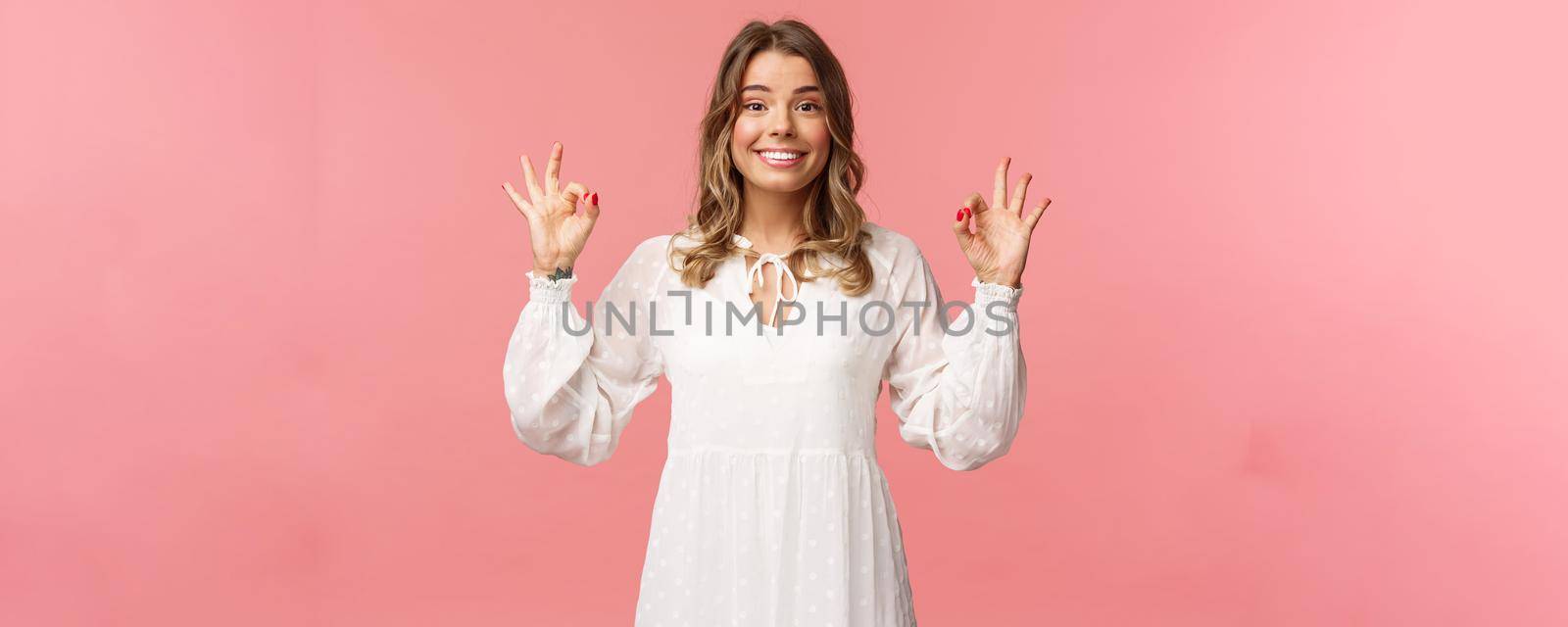 Beauty, fashion and women concept. Portrait of excited and upbeat blond female in white dress, standing satisfied over pink background, show okay sign, guarantee and assure in quality by Benzoix