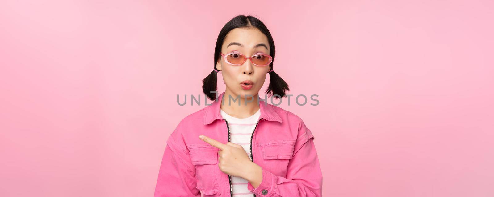 Close up portrait of asian woman gasping, looking surprised, pointing finger at banner, advertisement, standing over pink background.