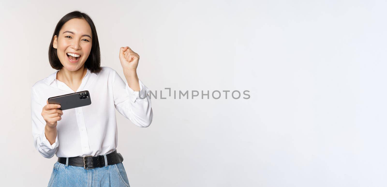 Happy asian woman do winner dance, triumphing, winning on mobile phone video game, holding smartphone horizontal position and celebrating, white background.