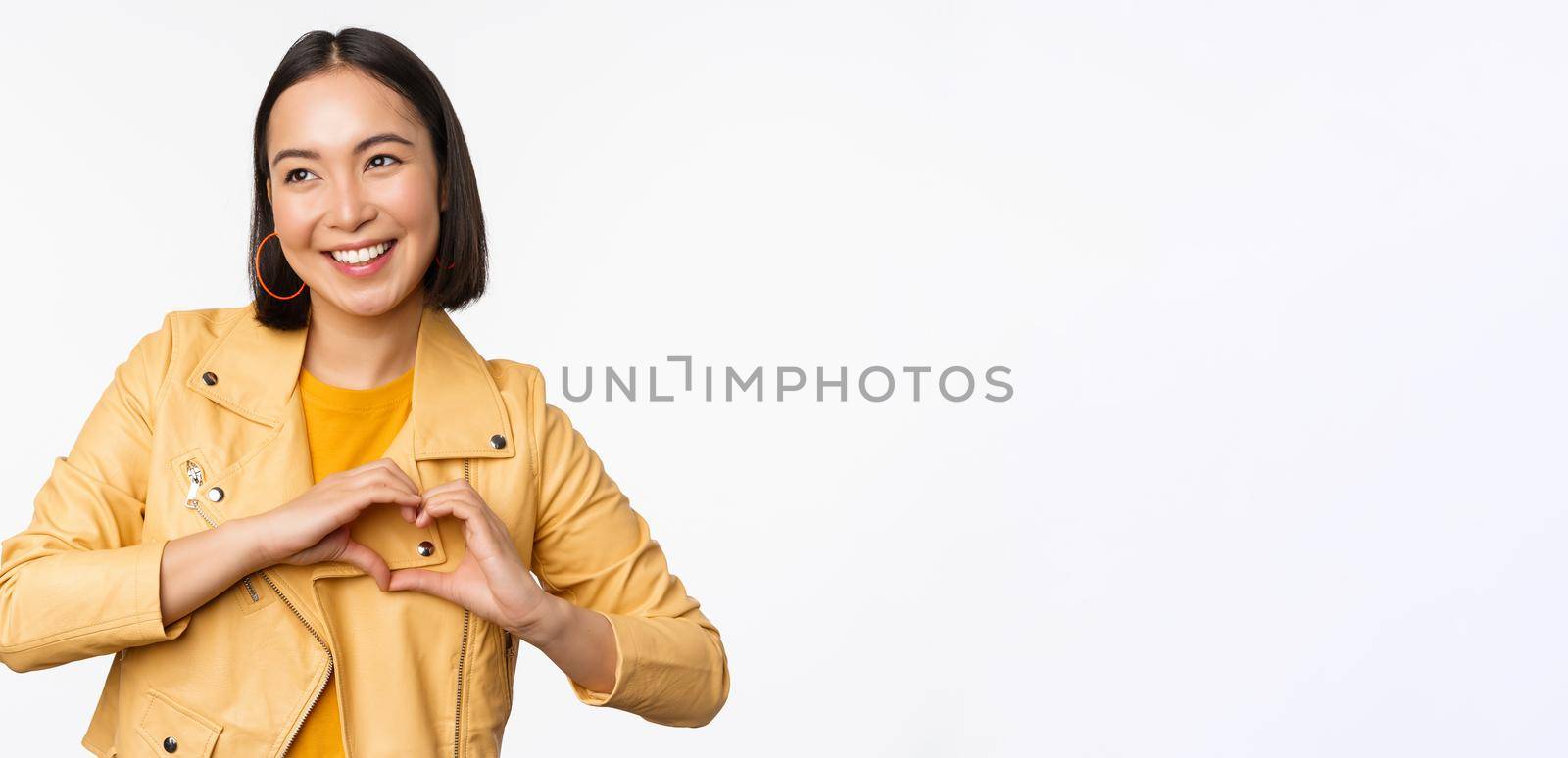 Image of beautiful asian girl showing heart, love sign, express care and romantic feelings, standing over white studio background.