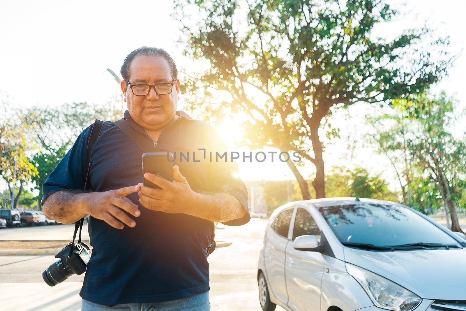 Local tourist mature Latin man using his cell phone and a camera hanging from his shoulder during sunset in a Nicaraguan outdoor park