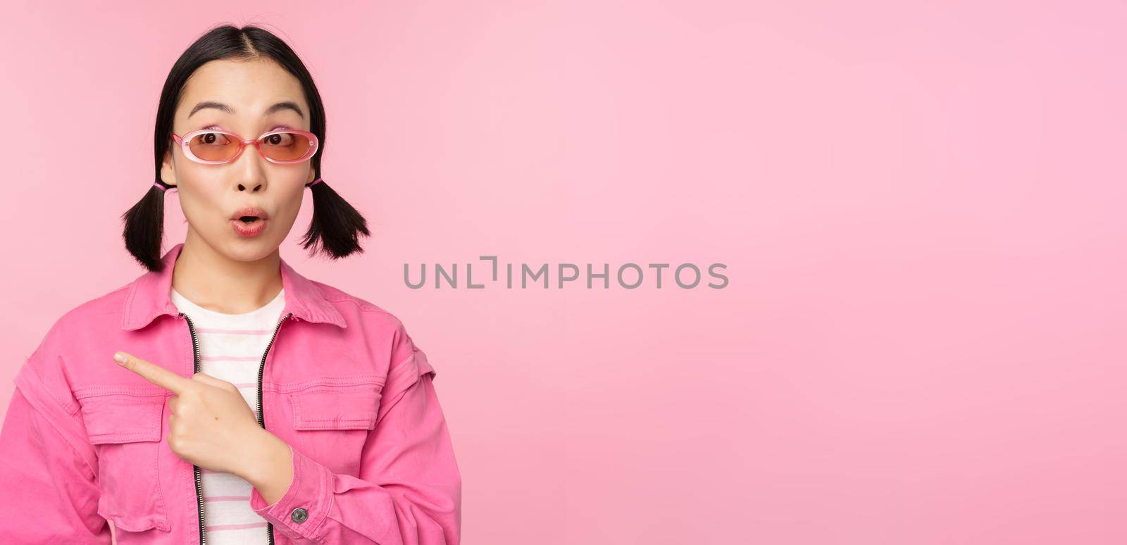 Portrait of smiling asian girl in stylish outfit, sunglasses, pointing finger left, showing advertisement, banner, standing over pink background. Copy space