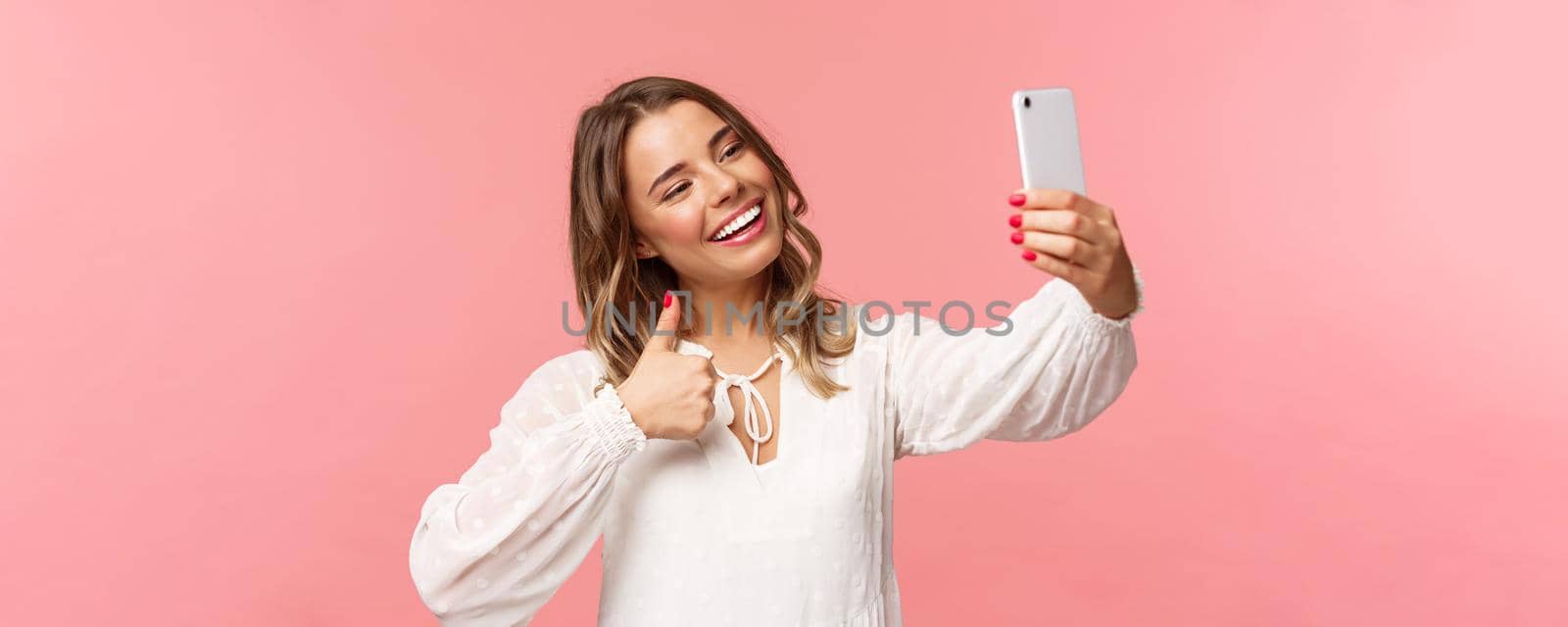 Close-up portrait of stylish beautiful beauty blogger record video on smartphone, taking selfie, showing thumbs-up at mobile phone camera, smiling pleased, recommend place to followers.