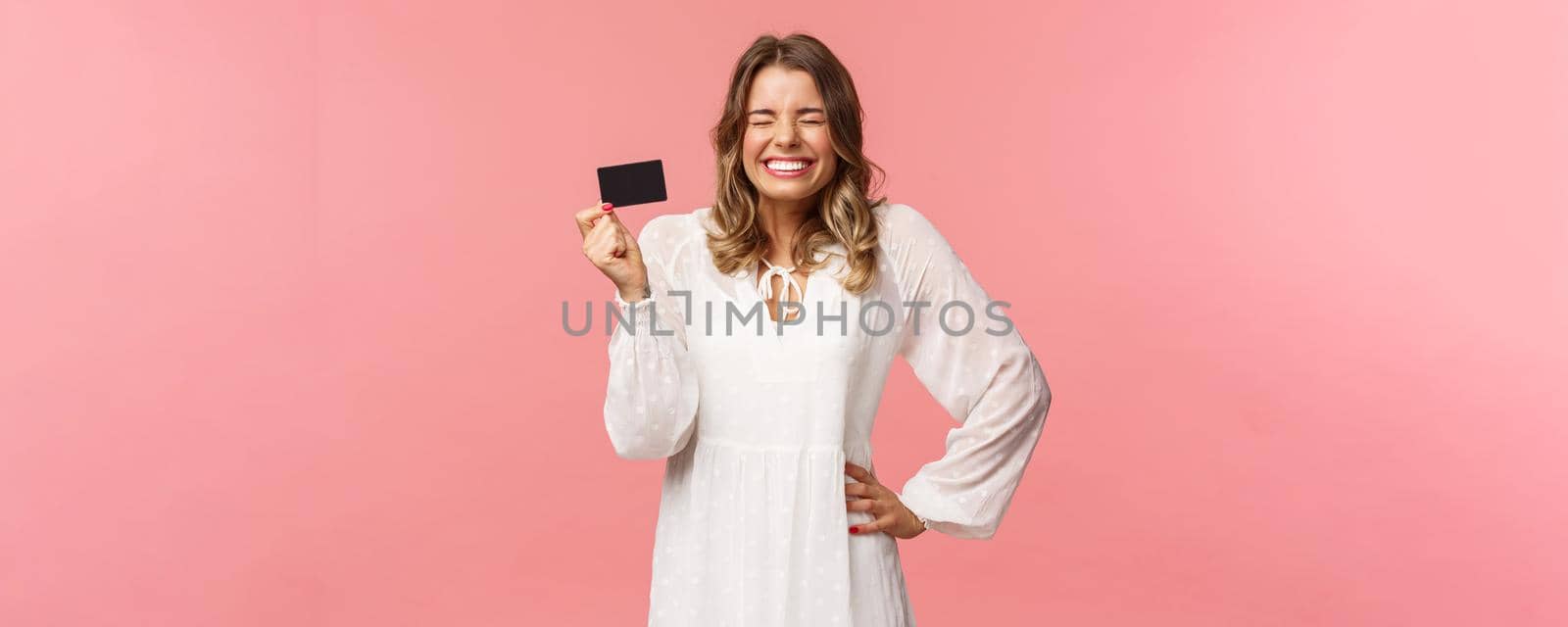 Portrait of excited cute and silly, feminine blond girl in white dress, close eyes giggle and smiling happy, got her first payment new job, holding credit card, use banking service, pink background by Benzoix