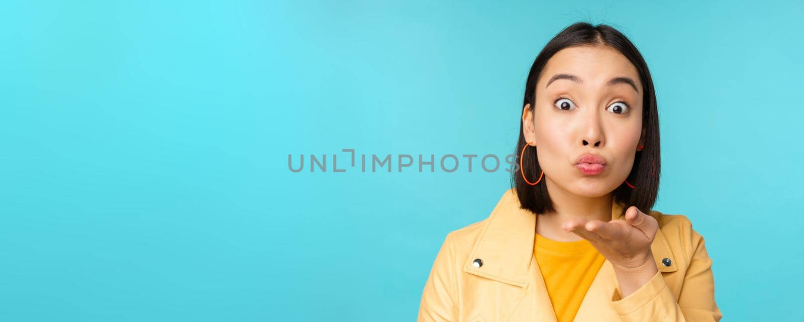 Close up portrait of funny asian girl sending air kiss, blowing at camera with popped eyes, standing over blue background.