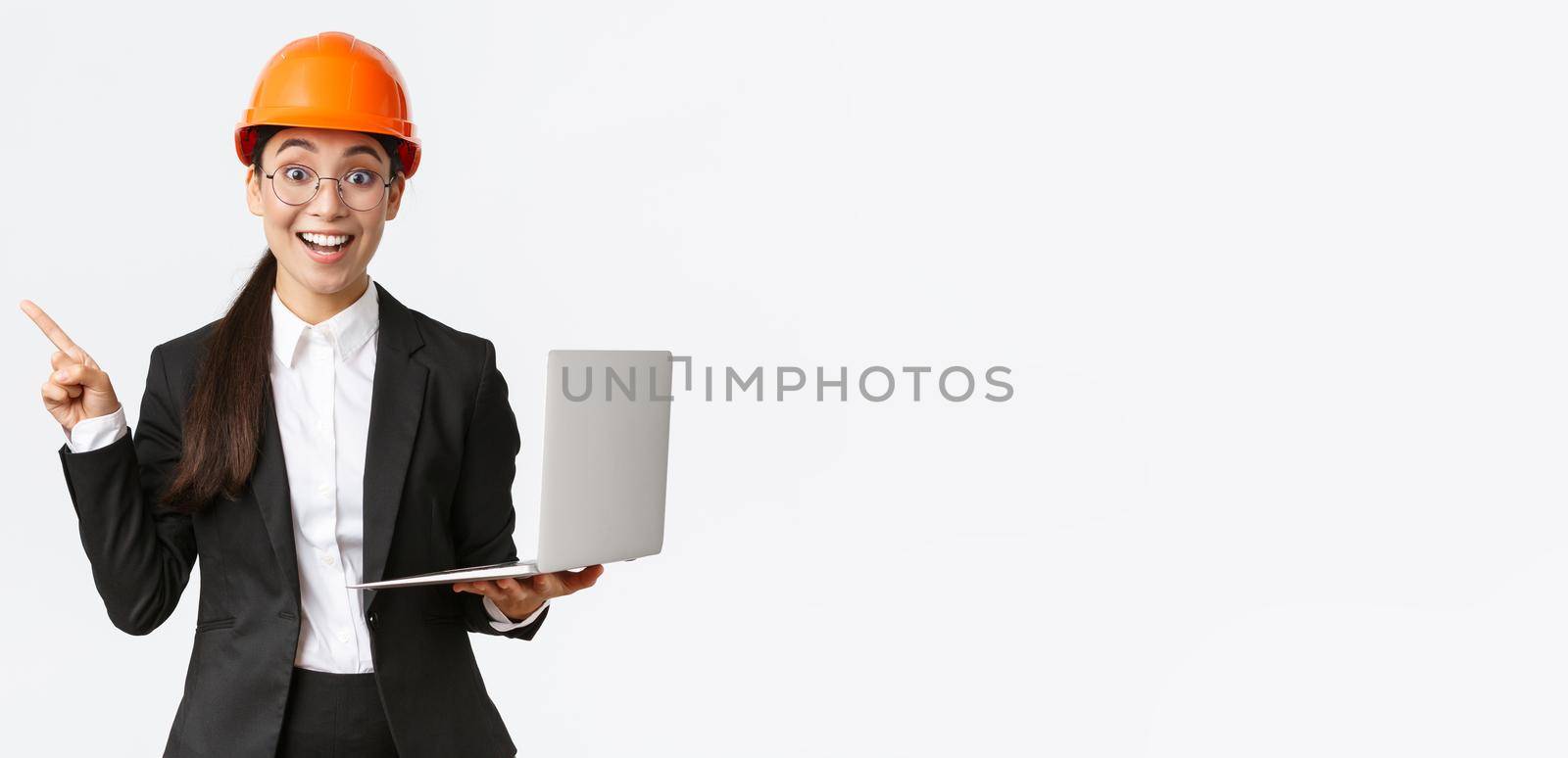Excited happy asian female engineer, industrial woman in safety helmet and business suit, showing presentation, pointing finger at graph or chart and holding laptop computer, smiling amazed.