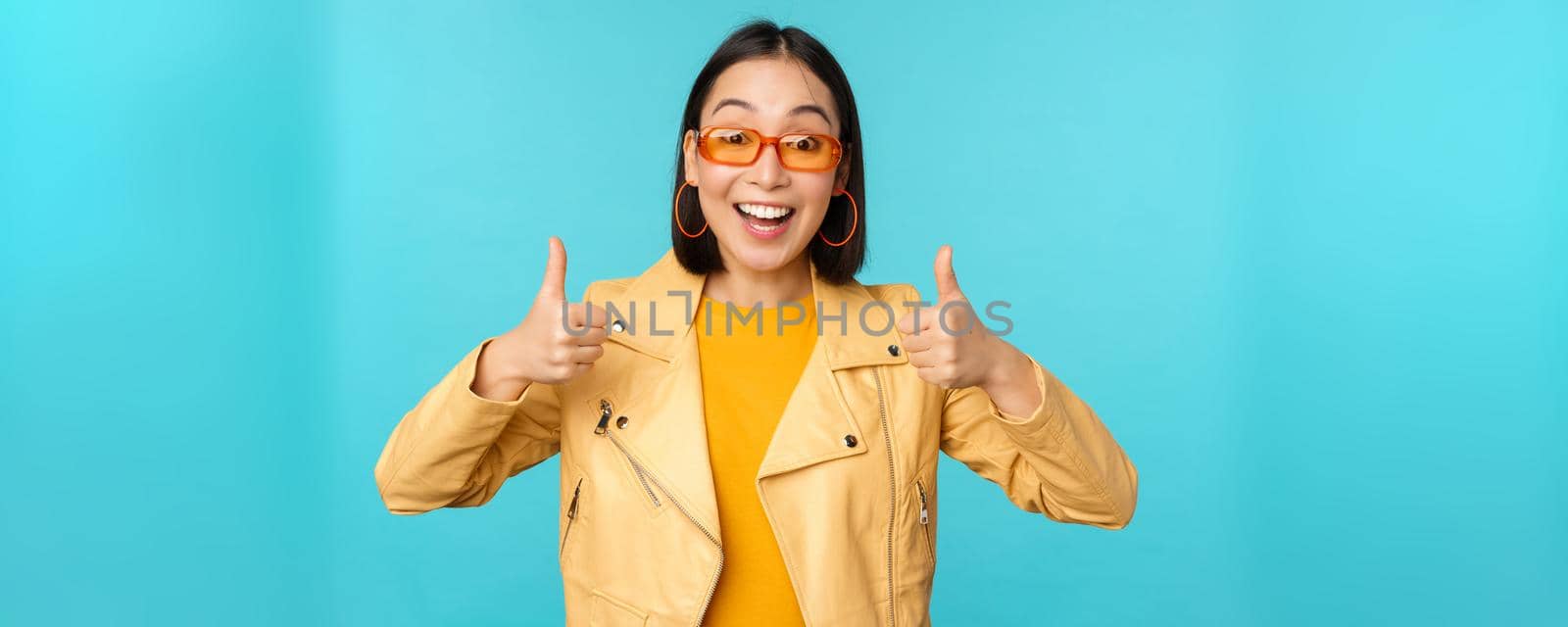 Enthusiastic korean girl looks excited and shows thumbs up, approves smth awesome, excellent choice and quality, stands over blue background.