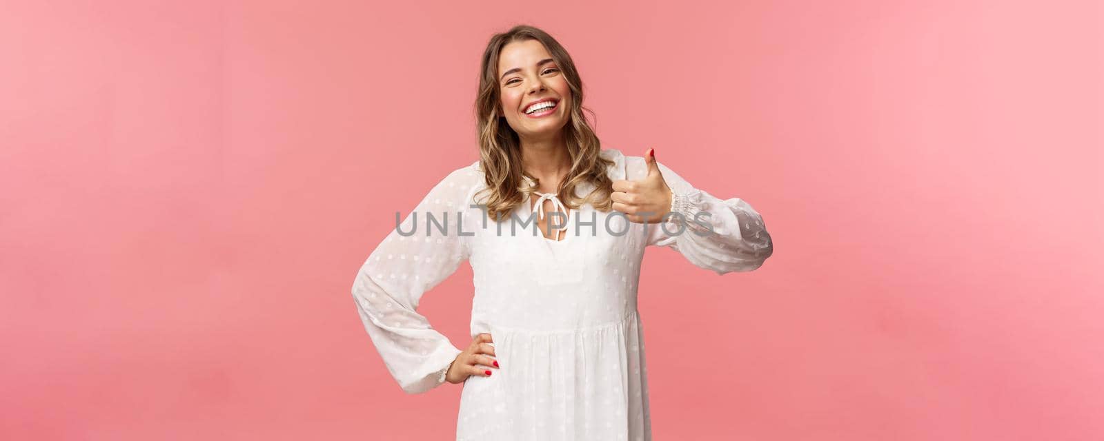 Enthusiastic lovely young woman in white cute dress, show thumb-up in approval, recommend product, smiling satisfied, leave positive review, impression, standing pink background.