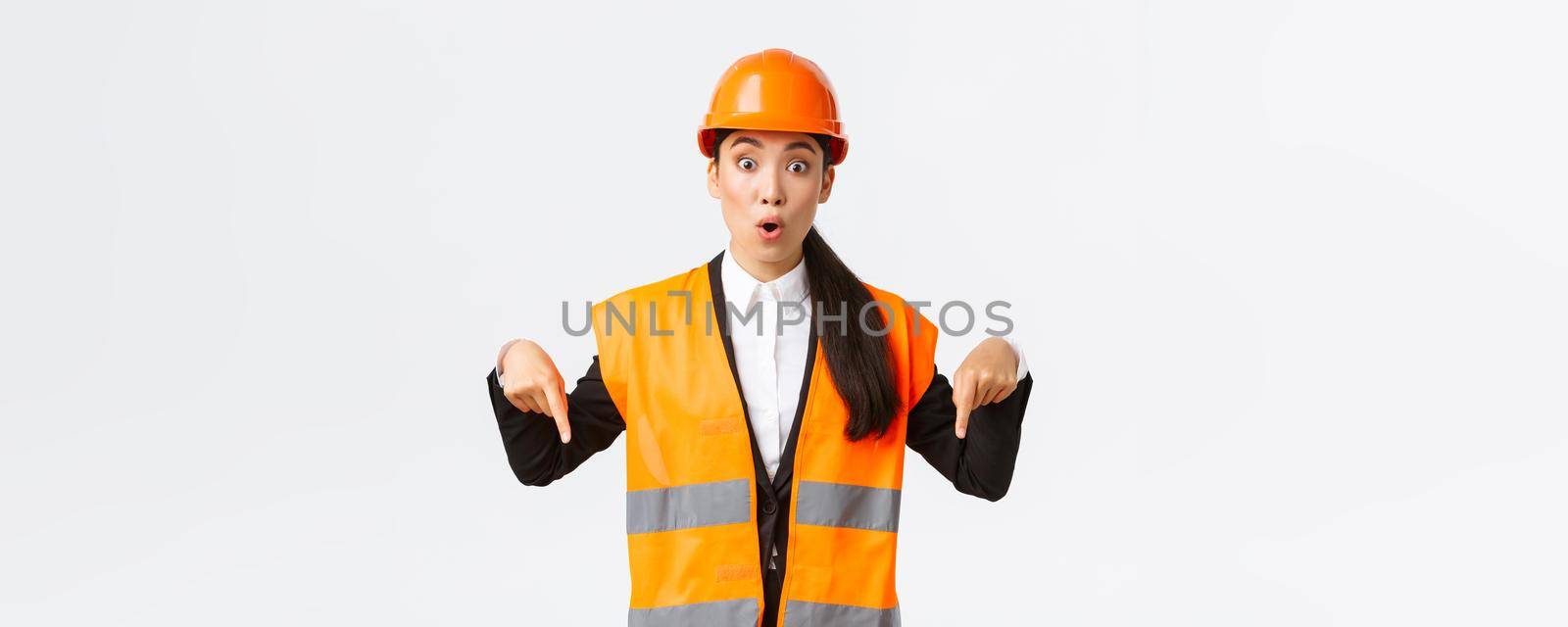 Amazed and interested asian female architect, industrial woman in reflective jacket, safety helmet pointing fingers down and looking impressed, talking about building construction, white background.