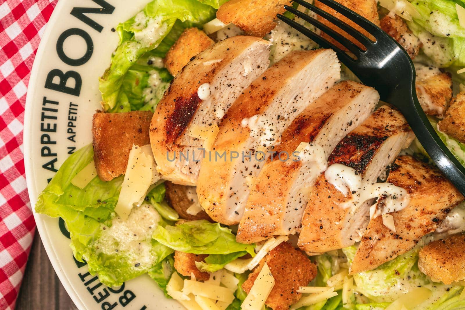 Caesar salad with thinly sliced chicken breast and traditional aioli on a plate that says bon apetite on a rustic table. Top view. Close-up detail. Natural food concept,