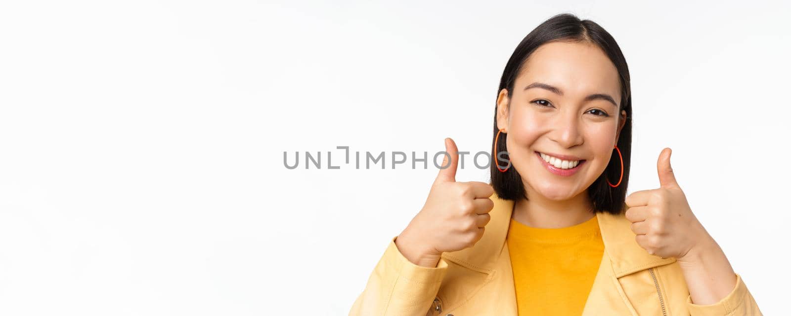 Smiling asian girl showing thumbs up, looking pleased, approve smth, standing over white background. Copy space