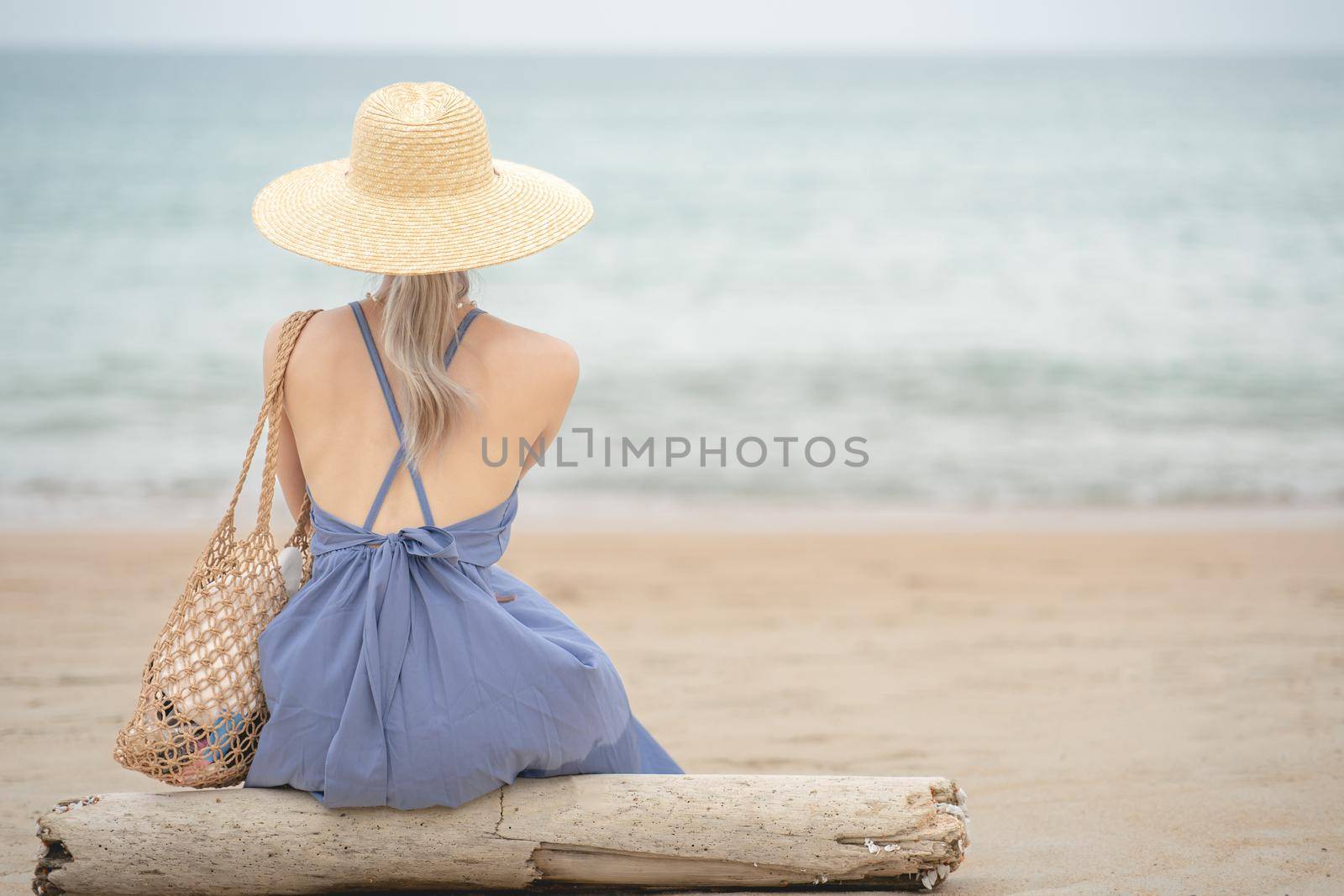 Woman in blue dress and straw hat, sitting on a timber by the ocean.
