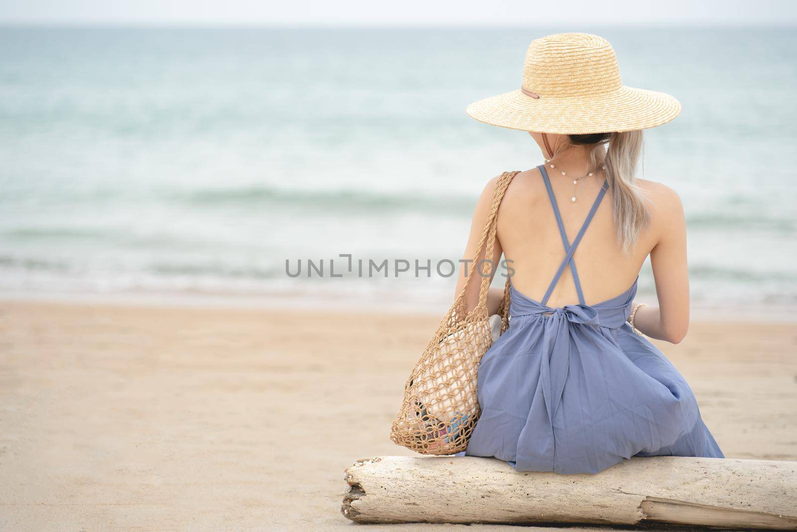 Woman in blue dress and straw hat, sitting on a timber by the ocean. by sirawit99