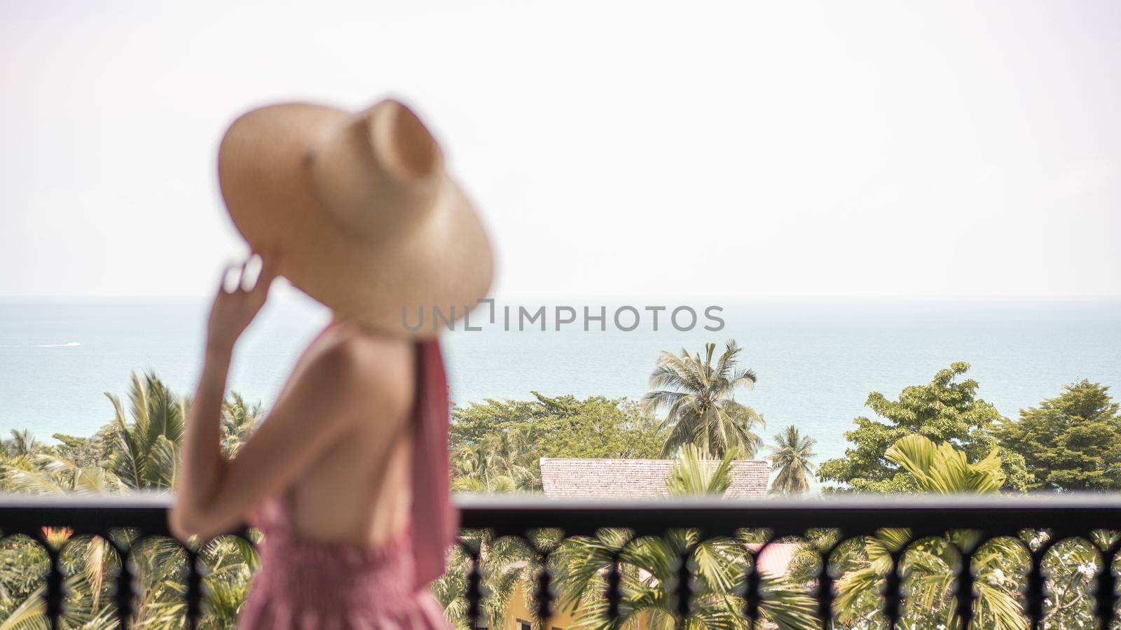 Blur side view of woman in pink dress and straw hat standing on a hotel balcony, ocean view. by sirawit99