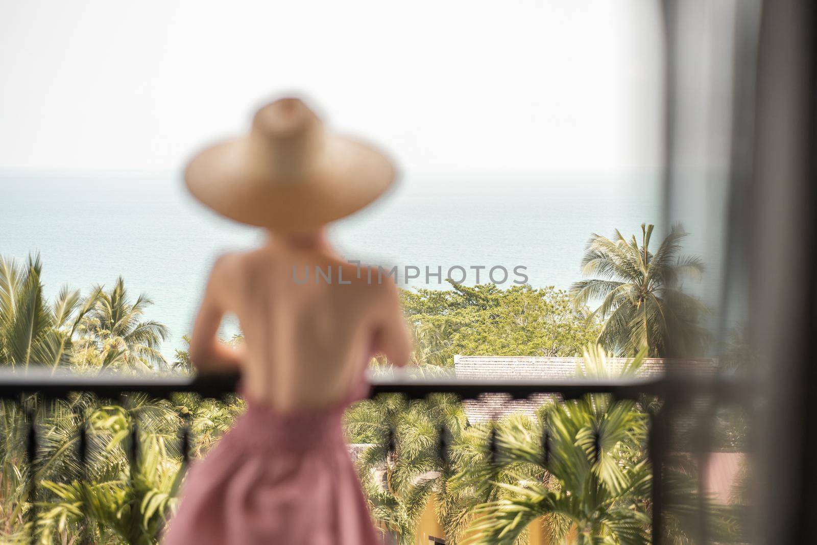 Blur back view of woman in pink dress and straw hat standing on a hotel balcony, ocean view. by sirawit99