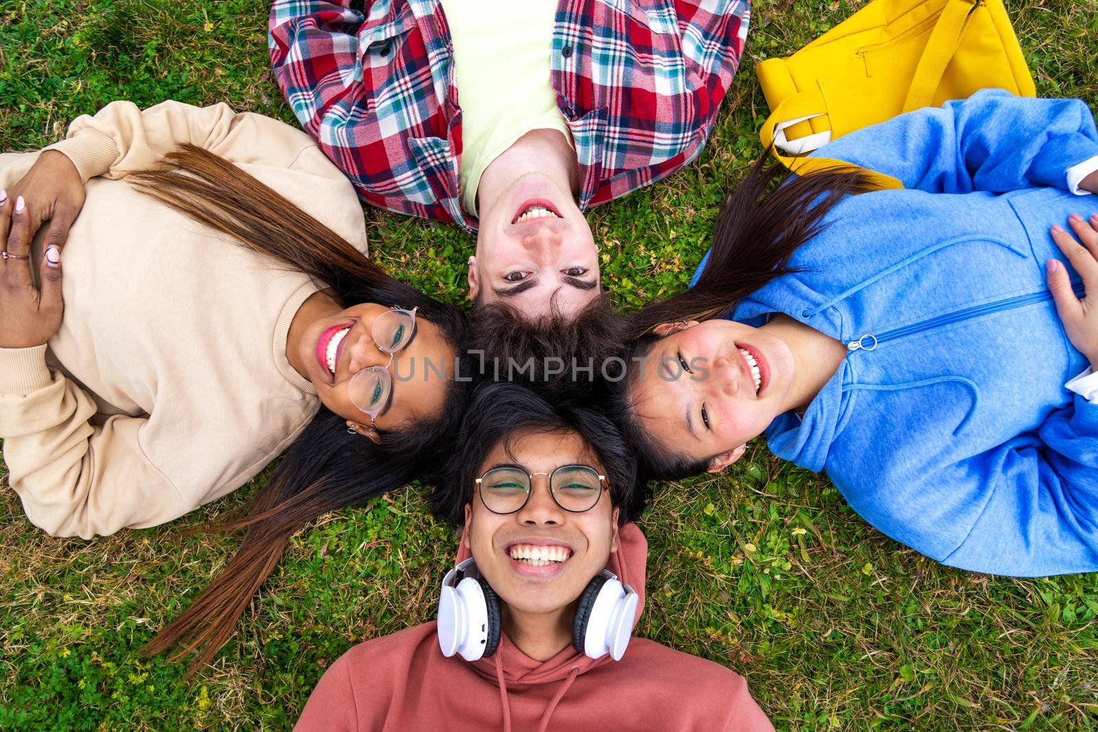 Group of happy and smiling multiracial college students lying on the grass looking at camera with heads in circle. Togetherness and relaxation concept.