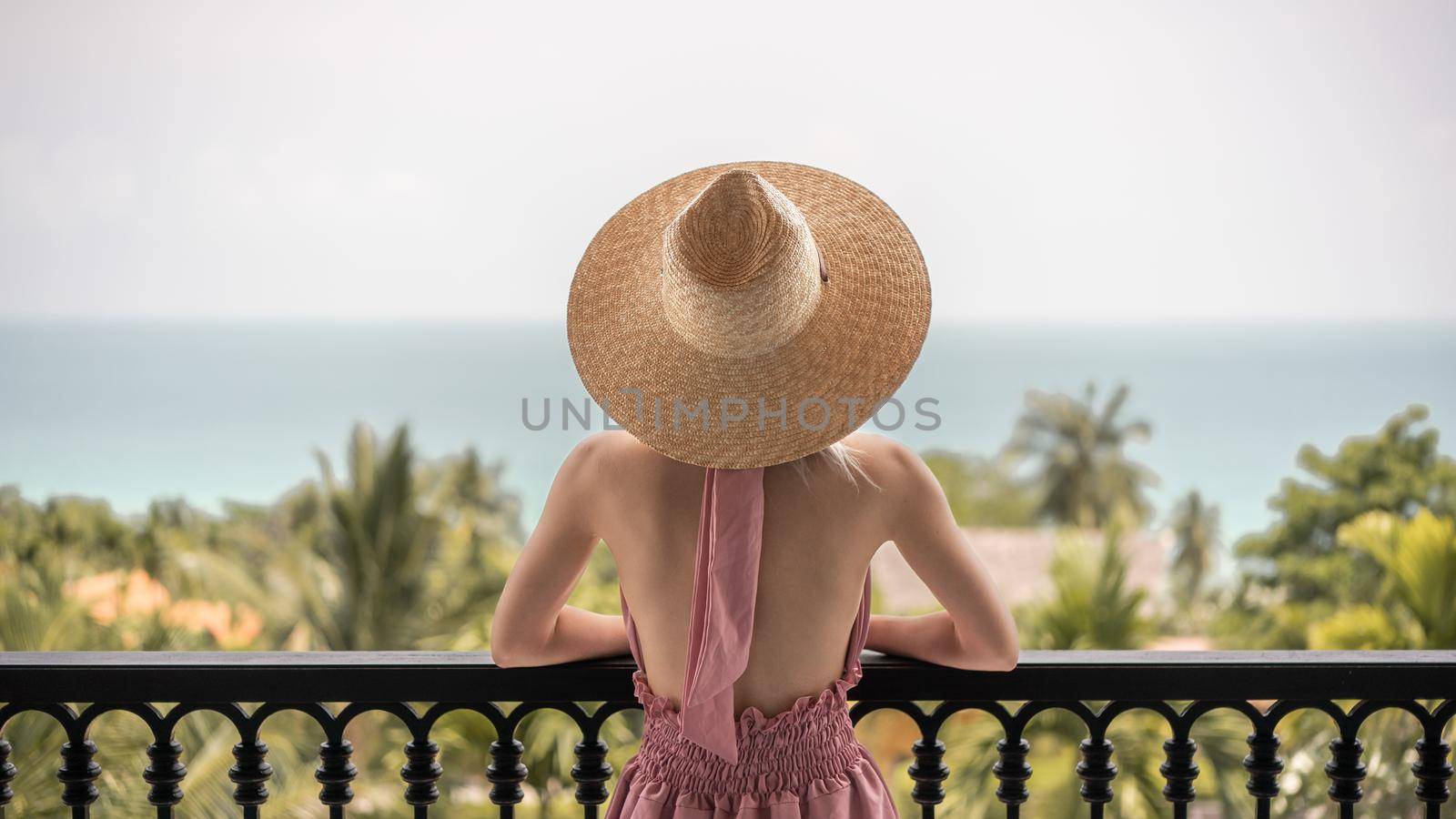 Back view of woman in pink dress and straw hat standing on a hotel balcony, ocean view. by sirawit99