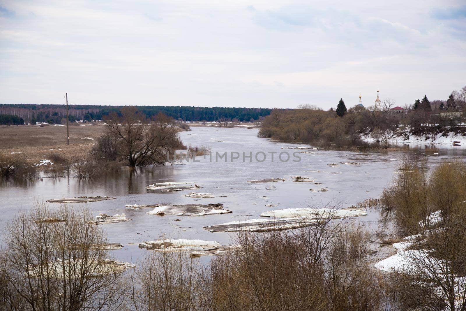 Small white ice floes float down the river. Spring, snow melts, dry grass all around, floods begin and the river overflows. Day, cloudy weather, soft warm light.