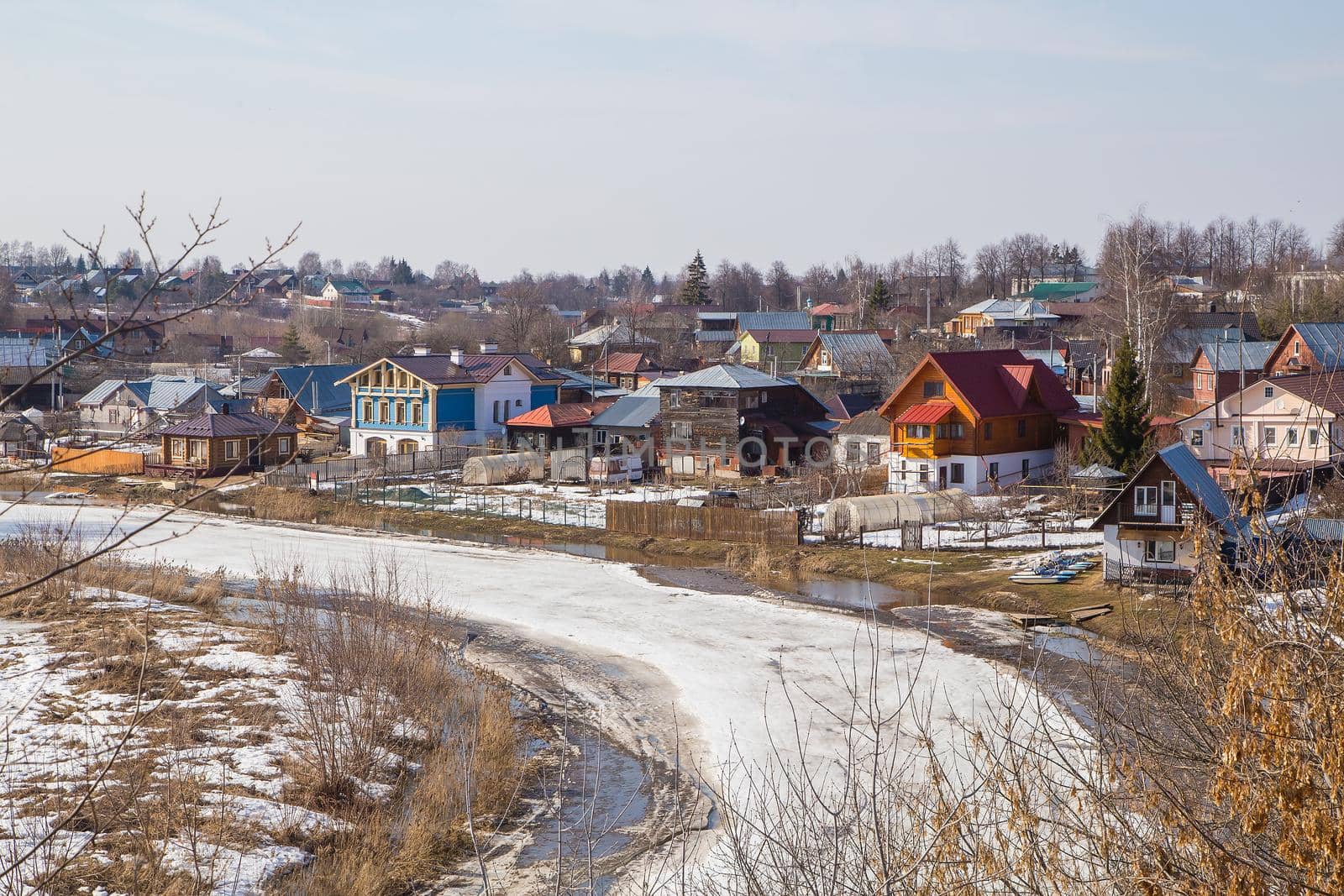 A frozen river stretching into the distance, a village on the bank. Spring, snow melts, puddles and dry grass all around. Day, cloudy weather, soft warm light.