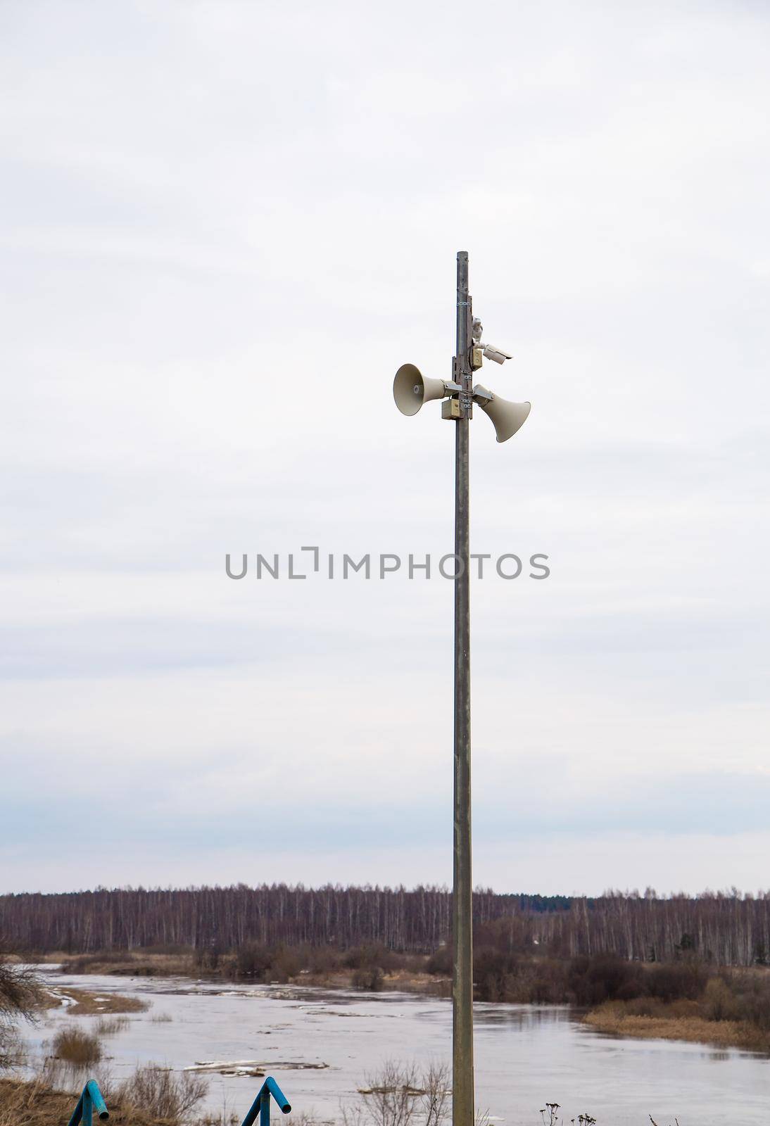Two loudspeakers-megaphones on a high pole against the background of the river. Spring, the snow is melting, there are puddles of slush and mud all around. Day, cloudy weather, soft warm light.