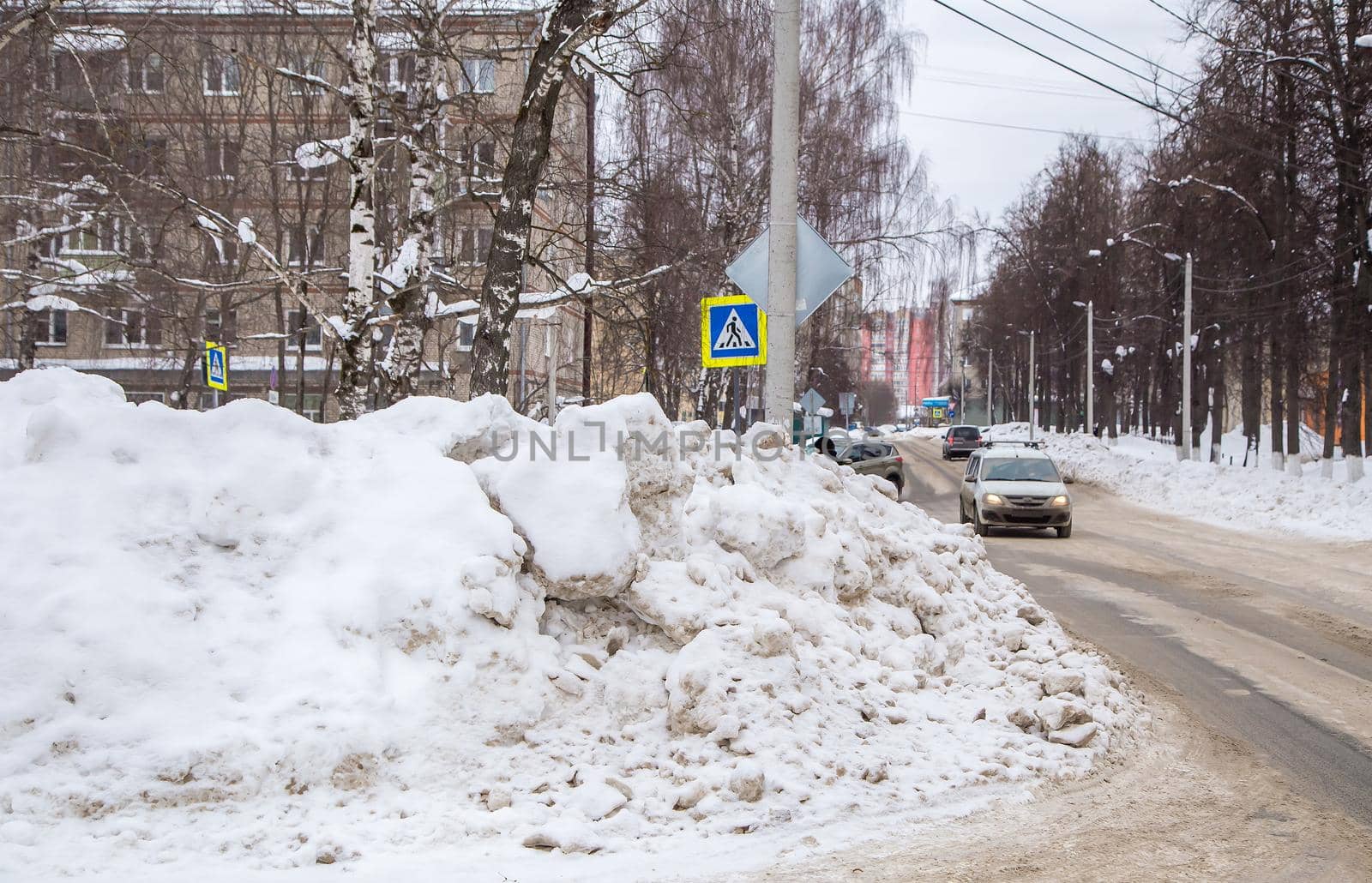 A huge large snowdrift by the road against the backdrop of a city street. On the road lies dirty snow in high heaps. Urban winter landscape. Cloudy winter day, soft light.